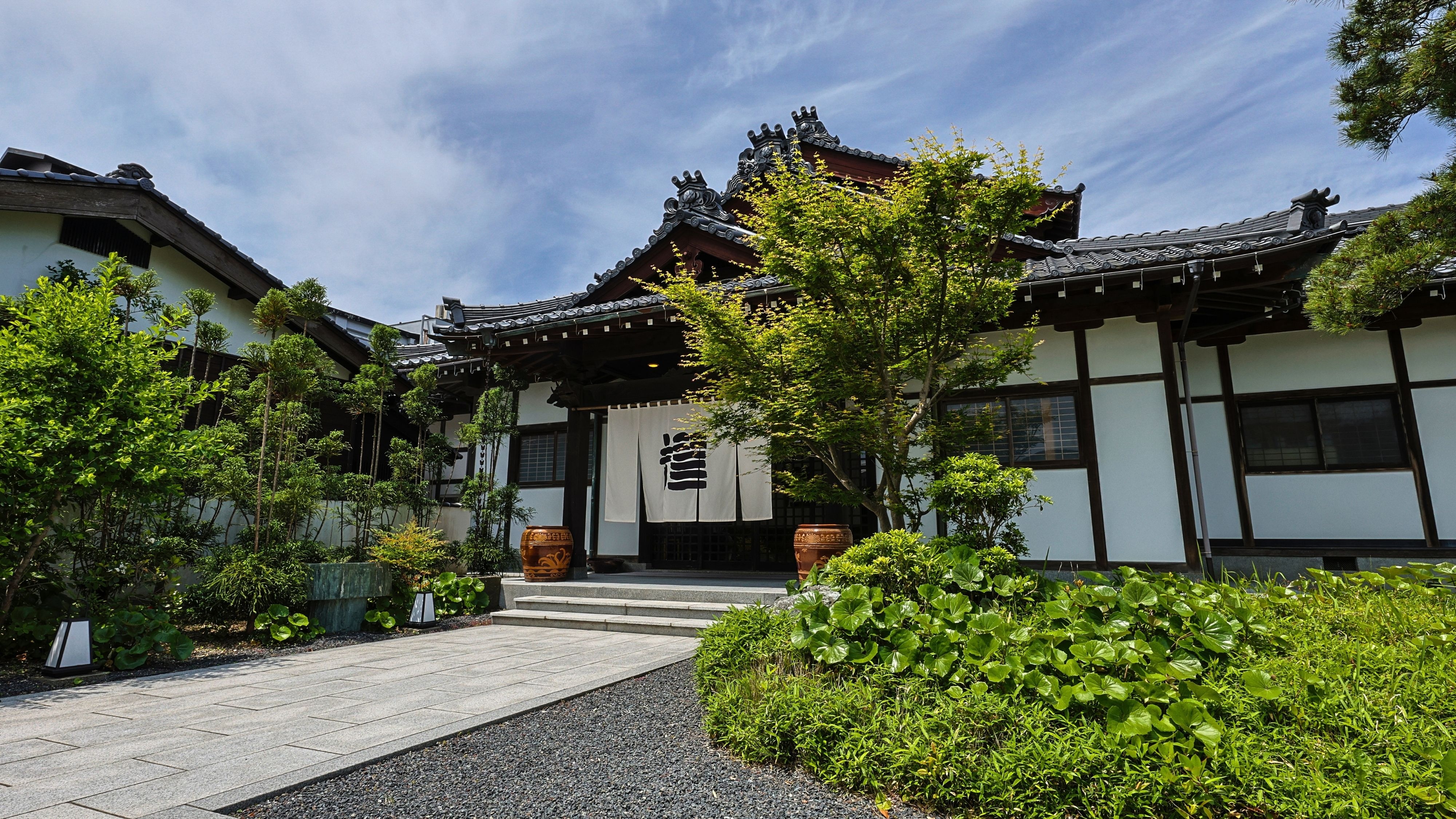 [Exterior] A noren (traditional Japanese curtain) beyond the dappled sunlight.